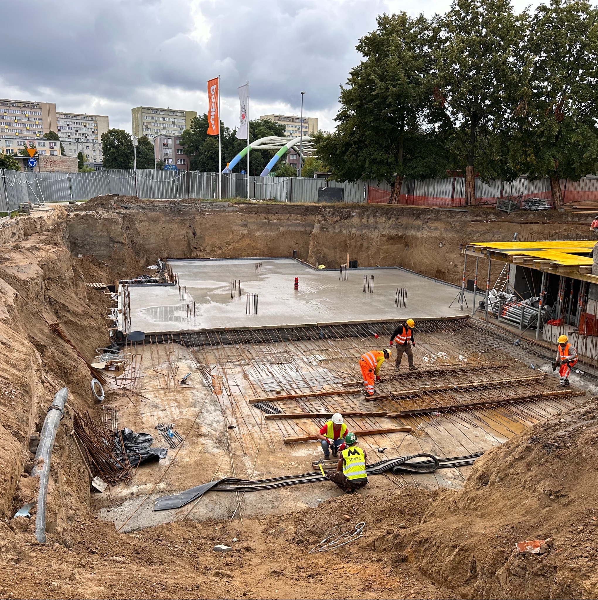 Zbrojenie fundamentów hali w wykopie, robotnicy w odblaskowych ubraniach układają stalowe pręty. W tle budynki mieszkalne i flaga firmy.