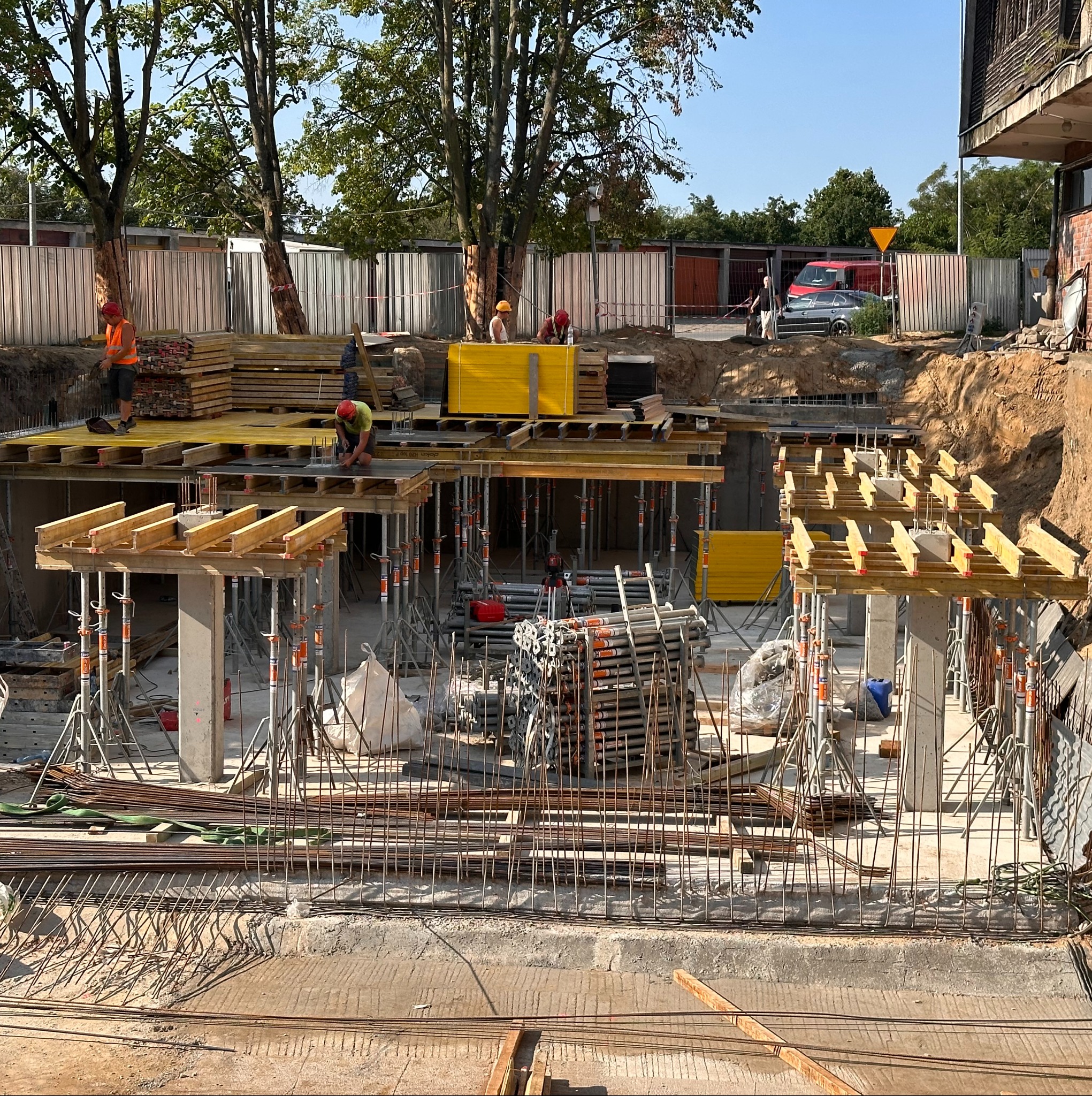 Fundamenty hali w trakcie budowy: zbrojenie, szalunki, podpory. Widoczni pracownicy w kaskach i kamizelkach. Na drugim planie ogrodzenie i budynki.