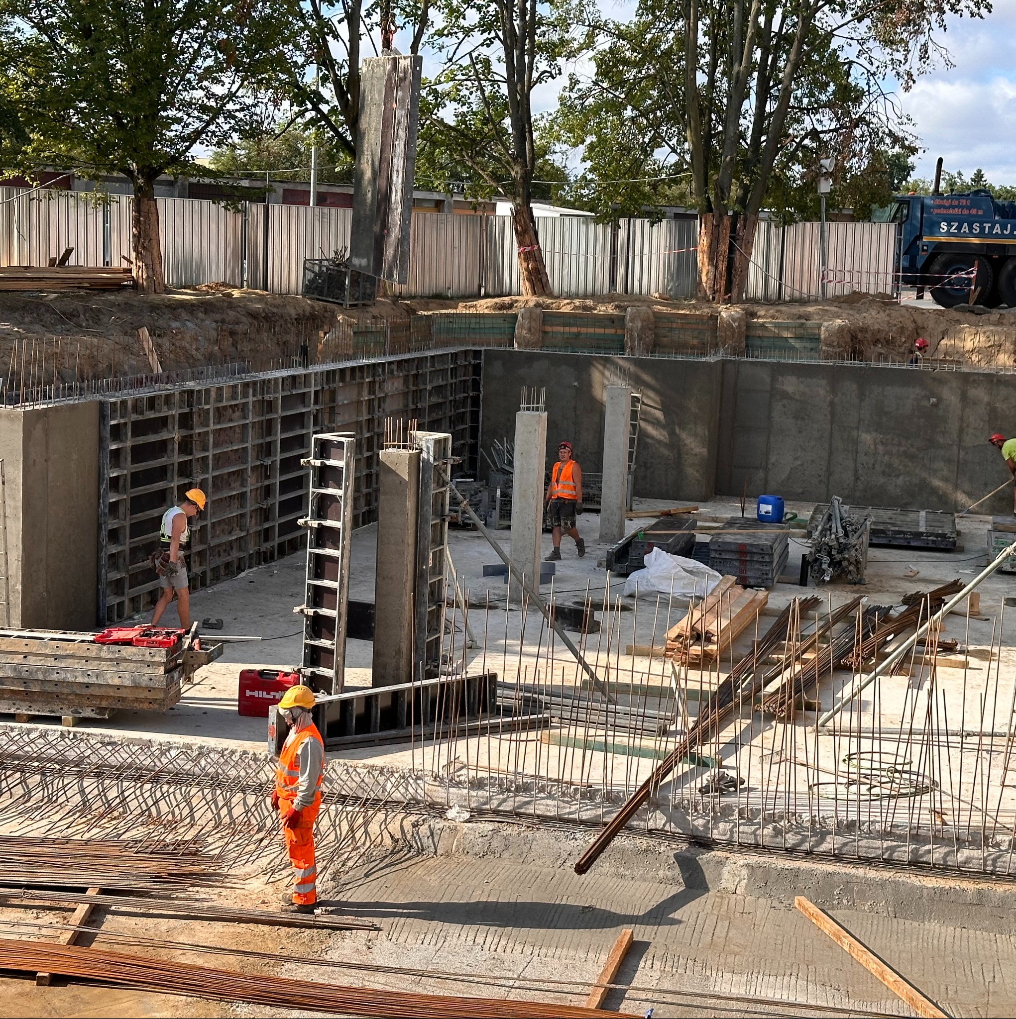 Fundamenty hali w trakcie budowy: zbrojenie, szalunki, betonowe ściany. Pracownicy w kaskach i kamizelkach odblaskowych na placu budowy. Widok z góry na teren budowy.