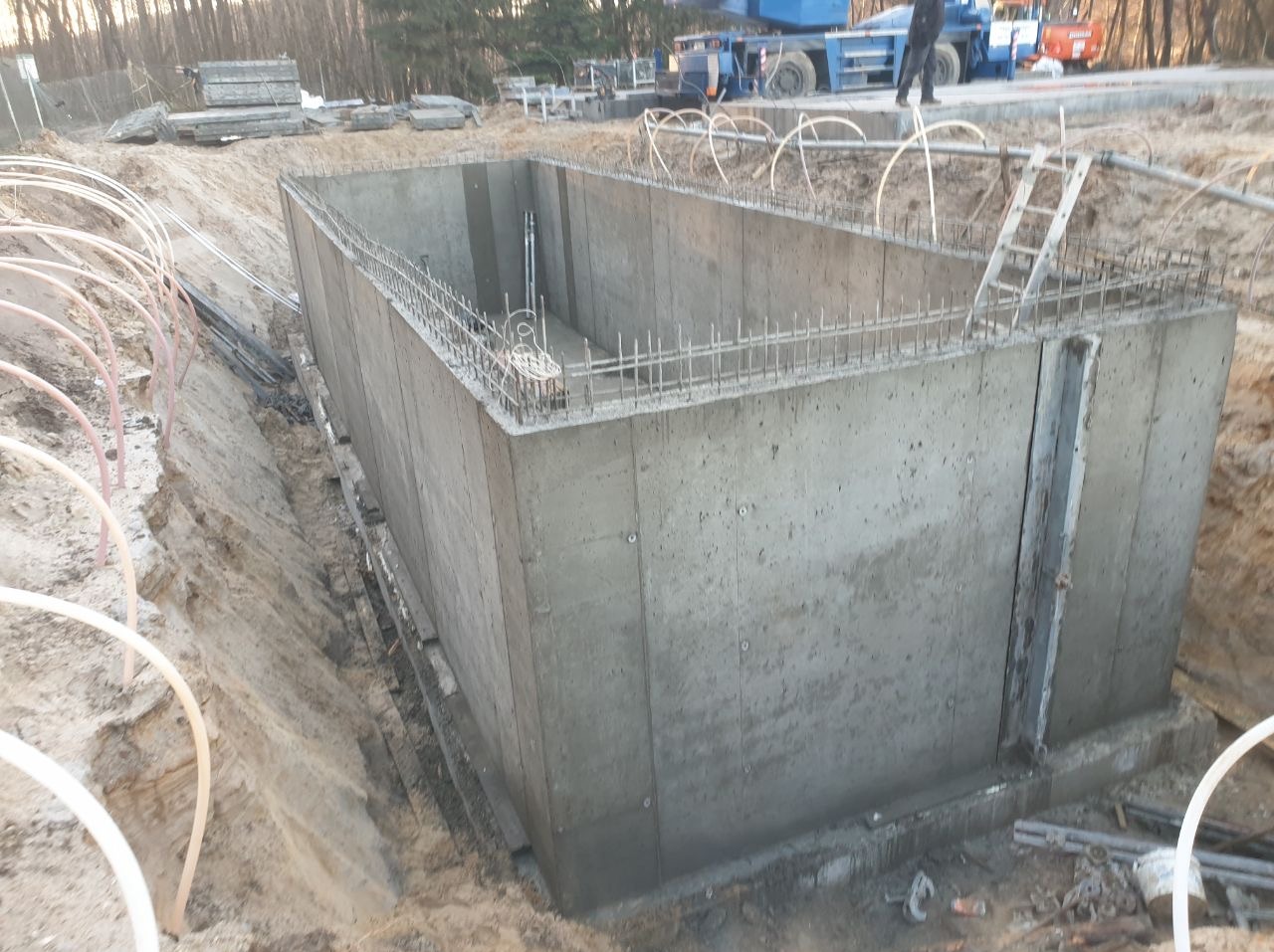 Betonowy fundament z wystającym zbrojeniem, otoczony wykopem i instalacjami. W tle widoczny dźwig i drzewa. Ujęcie z góry, podkreślające solidność konstrukcji.