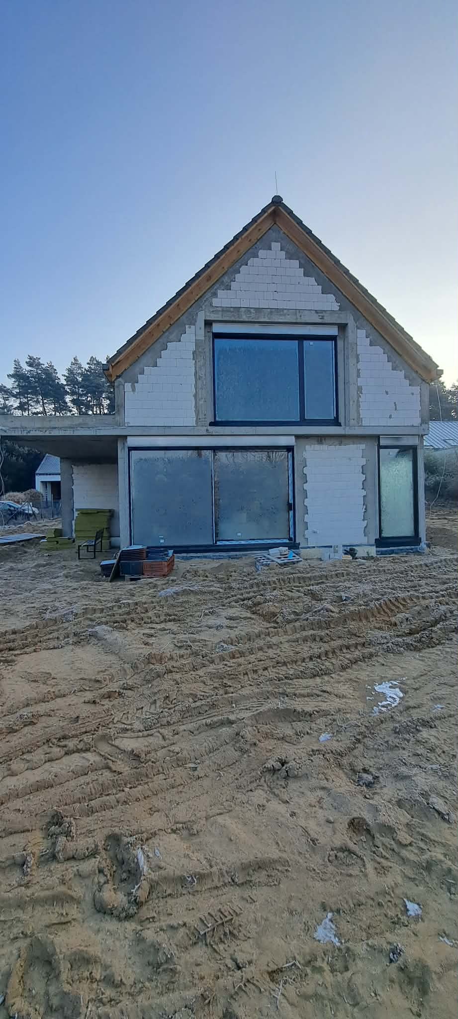 Budowa domu jednorodzinnego w stanie surowym otwartym. Widoczne bloczki, okna i przygotowany teren. Ujęcie z dołu, w tle drzewa i niebo.