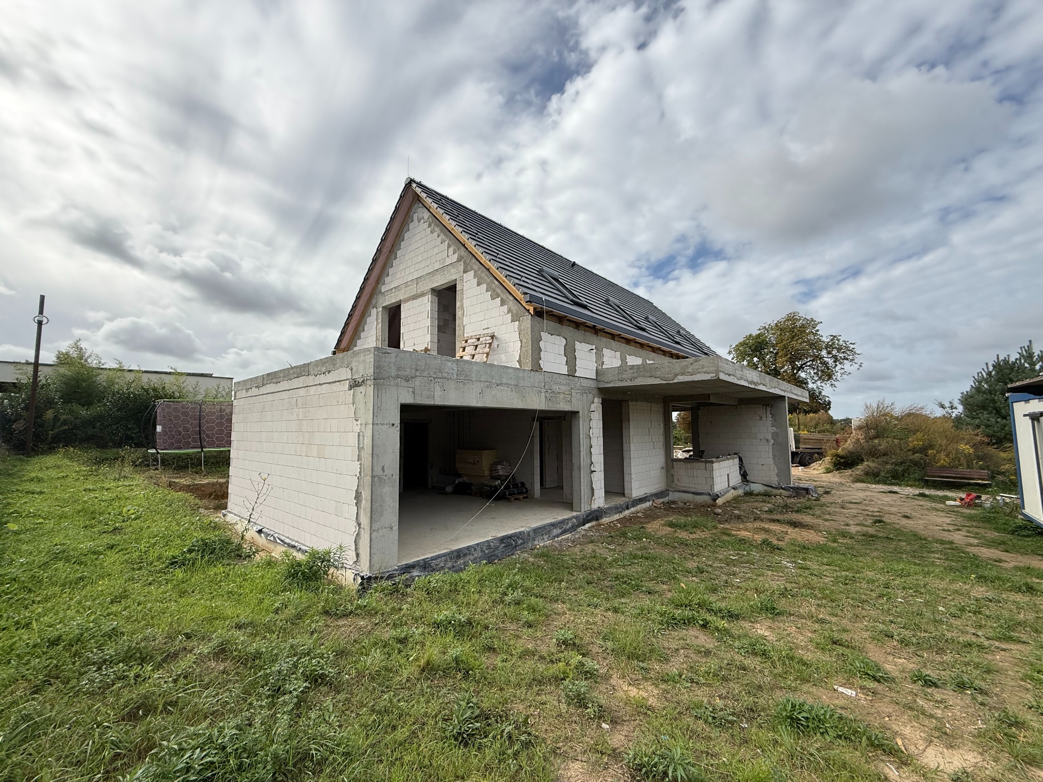 Budowa domu jednorodzinnego w stanie surowym otwartym z częściowo położonym dachem z dachówki, widok z zewnątrz na tle pochmurnego nieba.