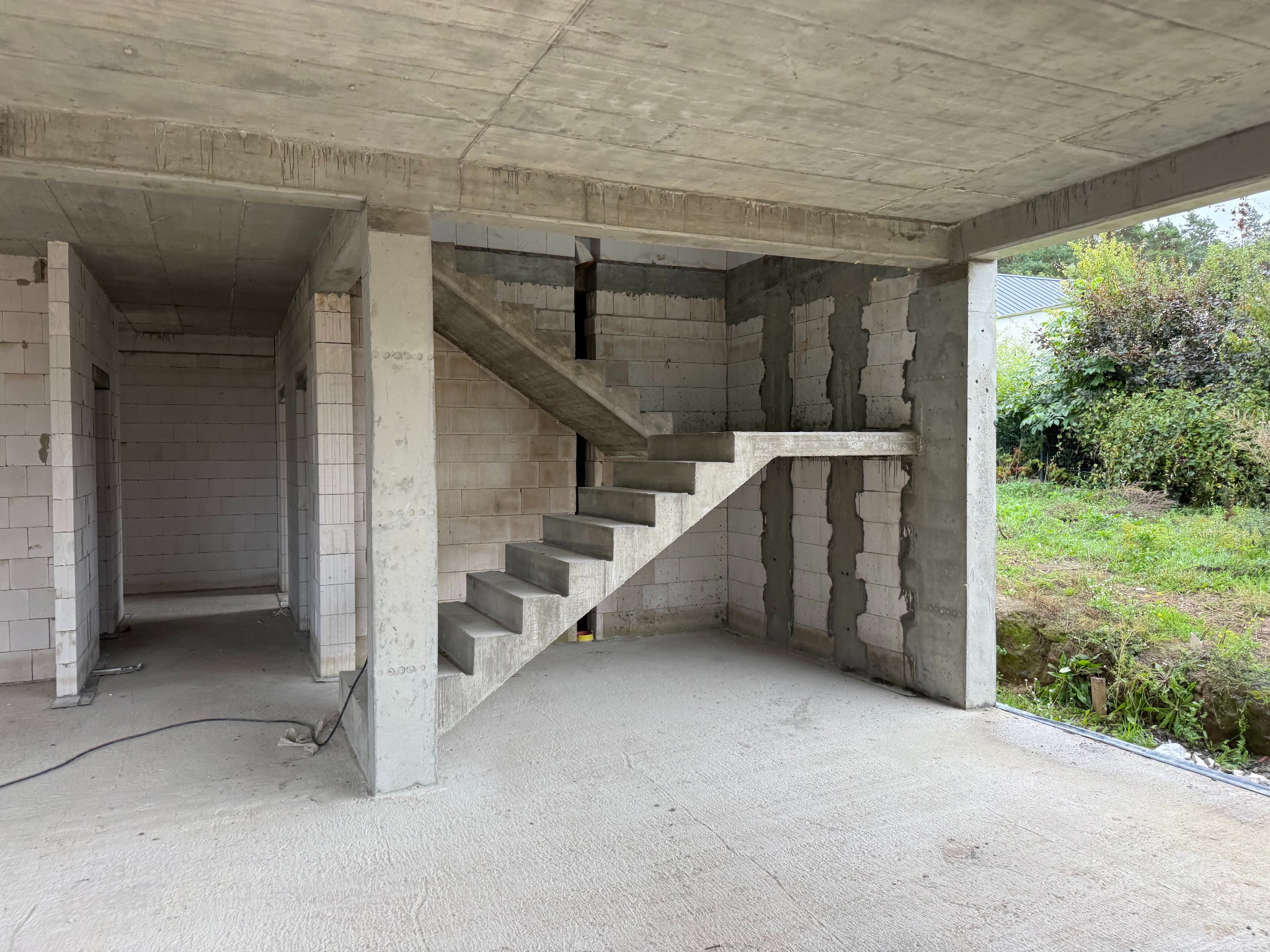 Surowe wnętrze hali w budowie: betonowe schody, ściany z białych bloczków, widoczne zbrojenia. Stan surowy otwarty, zieleń na zewnątrz.