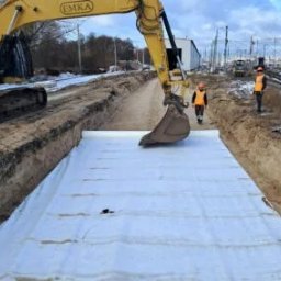 EMKA Kowalski Usługi Sp. z o.o. - Żółta koparka EMKA w wykopie wyłożonym białą geowłókniną, pracownicy w odblaskowych kamizelkach w tle, teren budowy w pobliżu torów kolejowych.