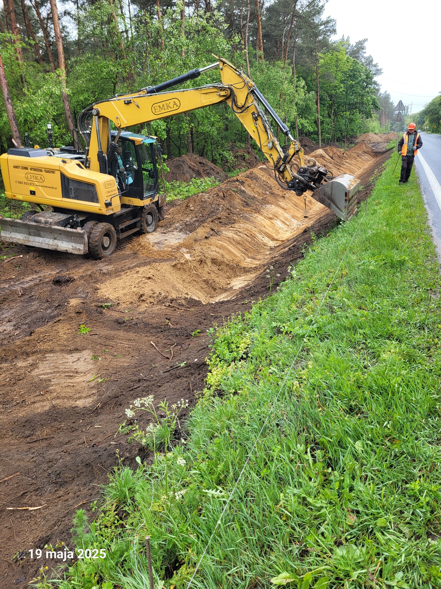 Żółta koparka EMKA wykonuje wykop rowu wzdłuż drogi, obok stoi pracownik w kamizelce odblaskowej. W tle las i zielona trawa. Widoczny napis '19 maja 2025'.