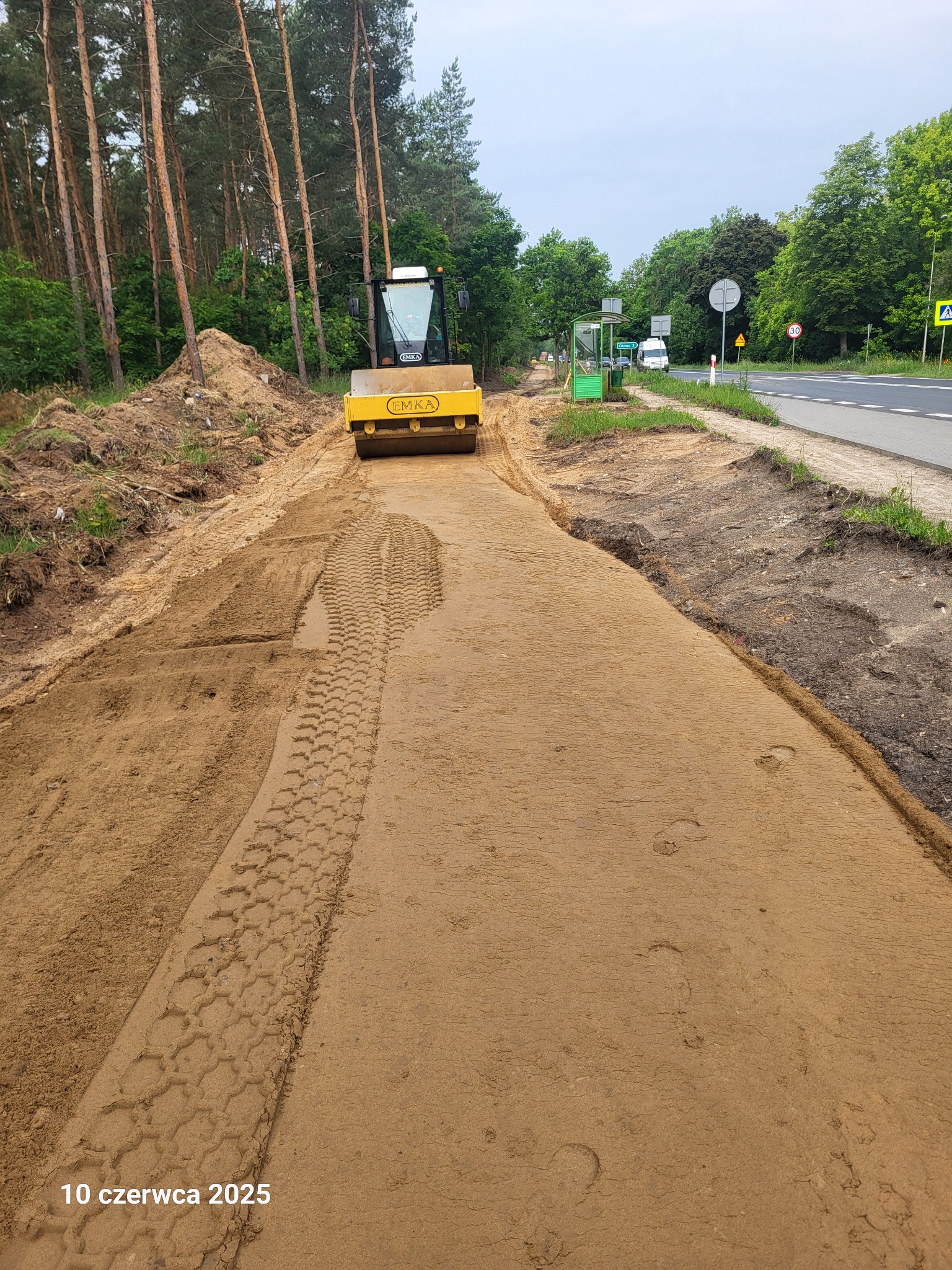 Żółty walec drogowy EMKA ubija piasek na nowo powstającej drodze. W tle las i droga publiczna z przystankiem autobusowym. Widoczne ślady opon walca.