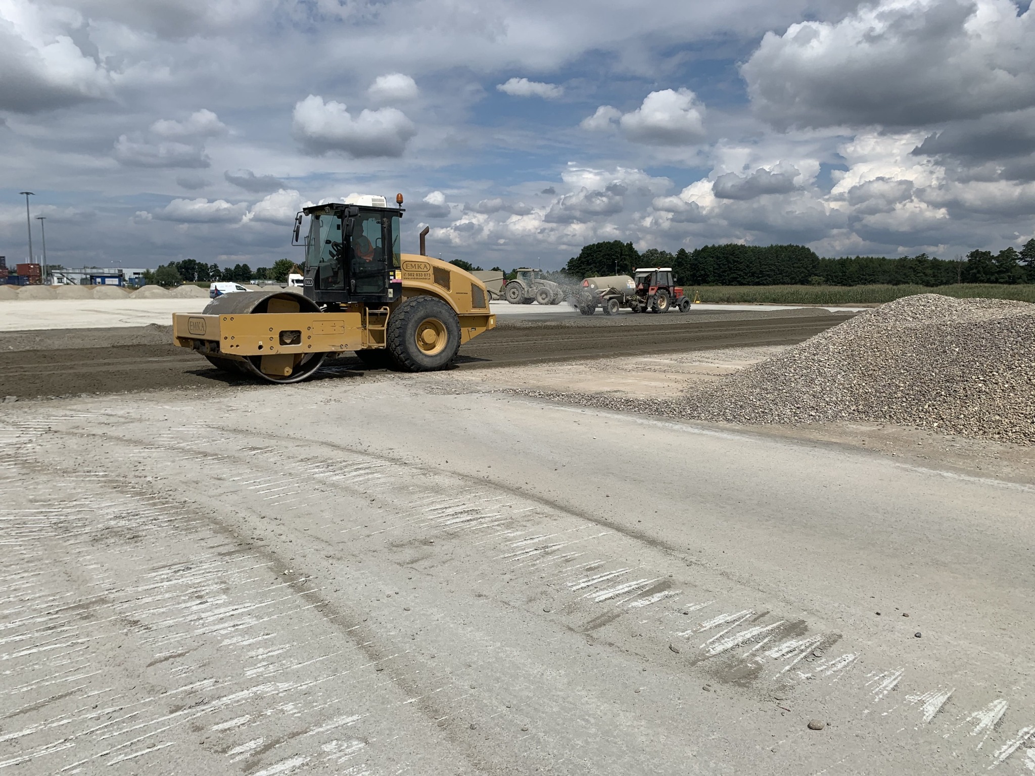 Żółty walec drogowy ubija szarą nawierzchnię. W tle widać traktory i hałdę żwiru pod pochmurnym niebem. Widoczne ślady po przejazdach maszyn.