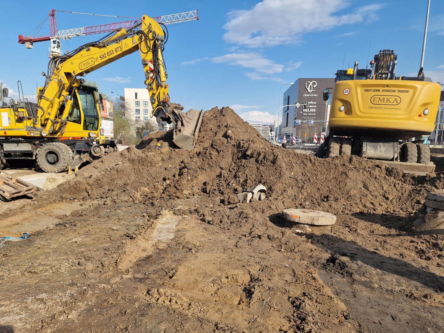Żółta koparka kołowa EMKA nabiera ziemię na placu budowy, w tle budynek handlowy i żuraw budowlany. Widoczne ślady opon na ziemi i elementy infrastruktury.