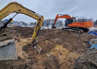 Dwa pracujące, żółto-pomarańczowe, ciężkie koparki w wykopie ziemnym, z betonowym kręgiem studni po lewej, na tle zabudowań miejskich w pochmurny dzień.