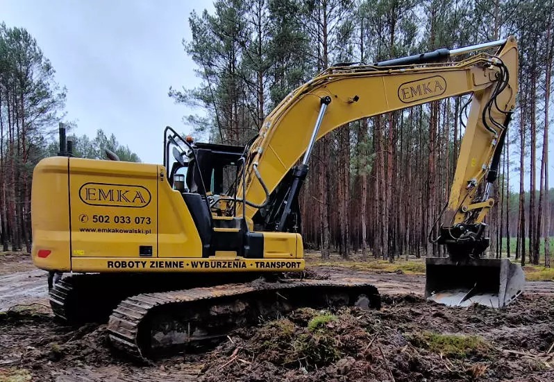 Żółta koparka EMKA na tle lasu sosnowego, z widocznymi śladami pracy w ziemi. Maszyna z łyżką zagłębia się w brudną ziemię, sugerując prace ziemne.