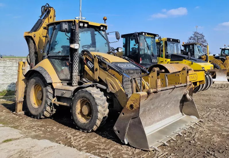 Żółta koparko-ładowarka 432F marki CAT na placu budowy, z widocznymi innymi maszynami w tle, w słoneczny dzień. Sprzęt budowlany gotowy do pracy.