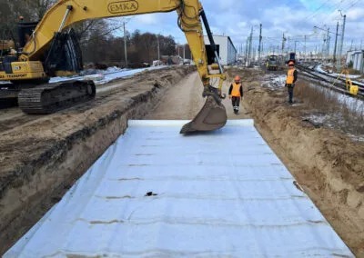 Żółta koparka EMKA w wykopie wyłożonym białą geowłókniną, pracownicy w odblaskowych kamizelkach w tle, teren budowy w pobliżu torów kolejowych.