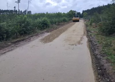Walec drogowy ubija świeżo wysypaną nawierzchnię drogi gruntowej, otoczoną zielenią i drzewami, pod pochmurnym niebem. Widok z perspektywy operatora.