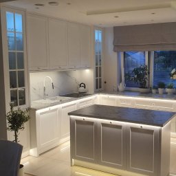 Sprzedaż Mebli Konrad Wójcik - Jasna kuchnia z wyspą, oświetleniem LED pod szafkami i marmurowym blatem. Okno z roletą rzymską i roślinami. Nowoczesny design z elementami klasycznymi.