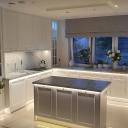 Sprzedaż Mebli Konrad Wójcik - Jasna kuchnia z wyspą, oświetleniem LED i marmurowym blatem. Okno z roletą rzymską i roślinami. Nowoczesny design z elementami klasycznymi.