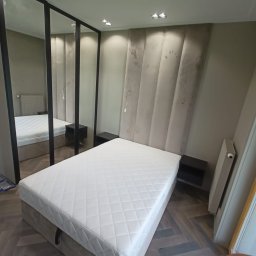 Sprzedaż Mebli Konrad Wójcik - Nowoczesna sypialnia z łóżkiem tapicerowanym, panelem ściennym i szafą z lustrzanymi drzwiami w czarnych ramach. Parkiet ułożony w jodełkę. Minimalistyczny design.