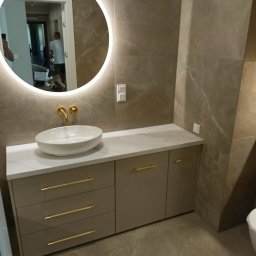 Sprzedaż Mebli Konrad Wójcik - Nowoczesna łazienka z szafką na wymiar w odcieniach beżu, z białym blatem i złotymi uchwytami. Okrągłe lustro z podświetleniem LED. Płytki imitujące marmur.