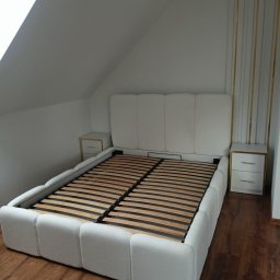 Sprzedaż Mebli Konrad Wójcik - Białe łóżko tapicerowane z drewnianym stelażem i szafkami nocnymi w pokoju na poddaszu. Wezgłowie z panelami i złotymi paskami. Podłoga z paneli.