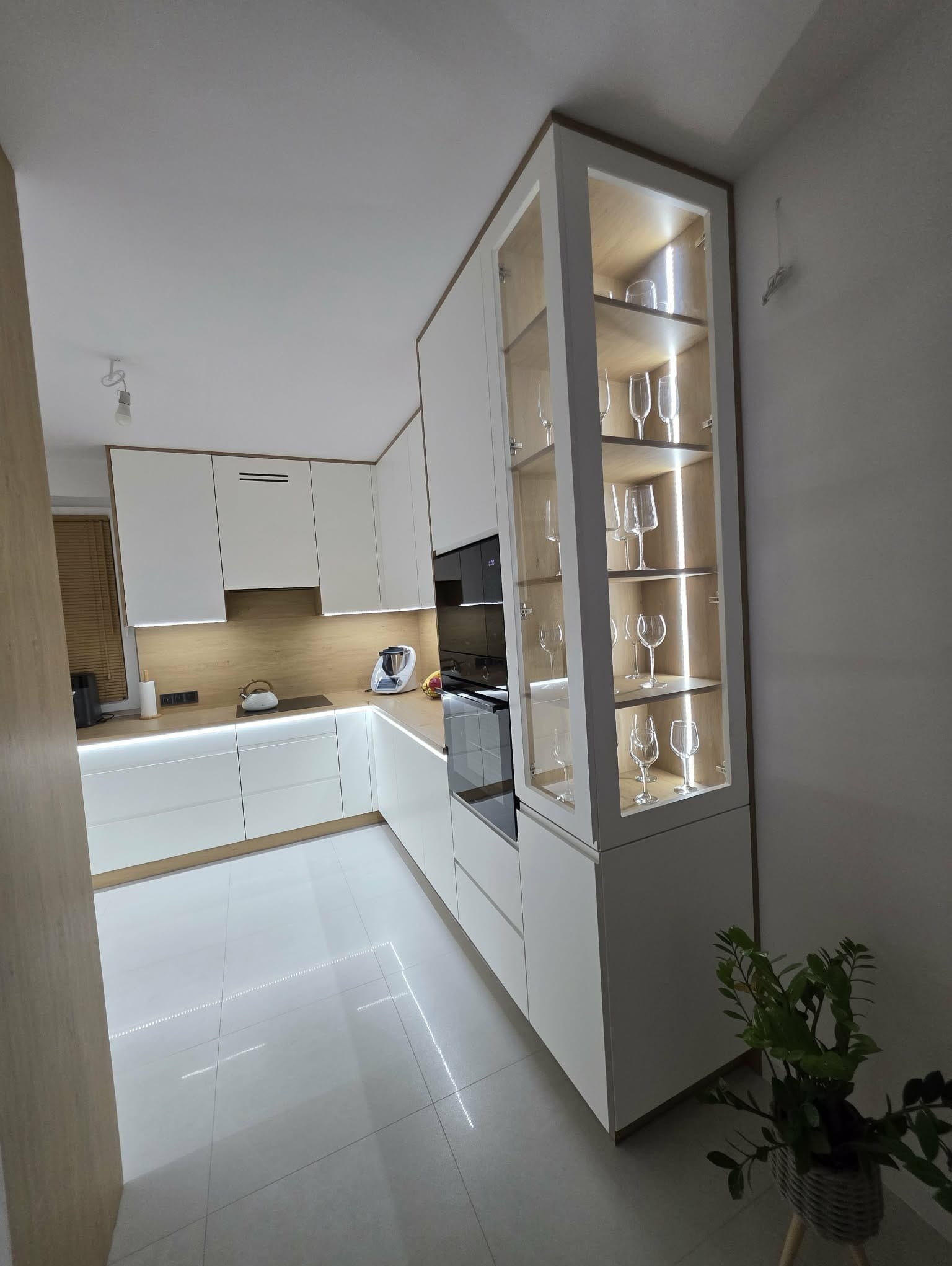 Nowoczesna, jasna kuchnia z białymi frontami i drewnianym blatem. Wysoka szafka z przeszklonymi drzwiami i podświetleniem eksponuje kieliszki. Minimalistyczny design.