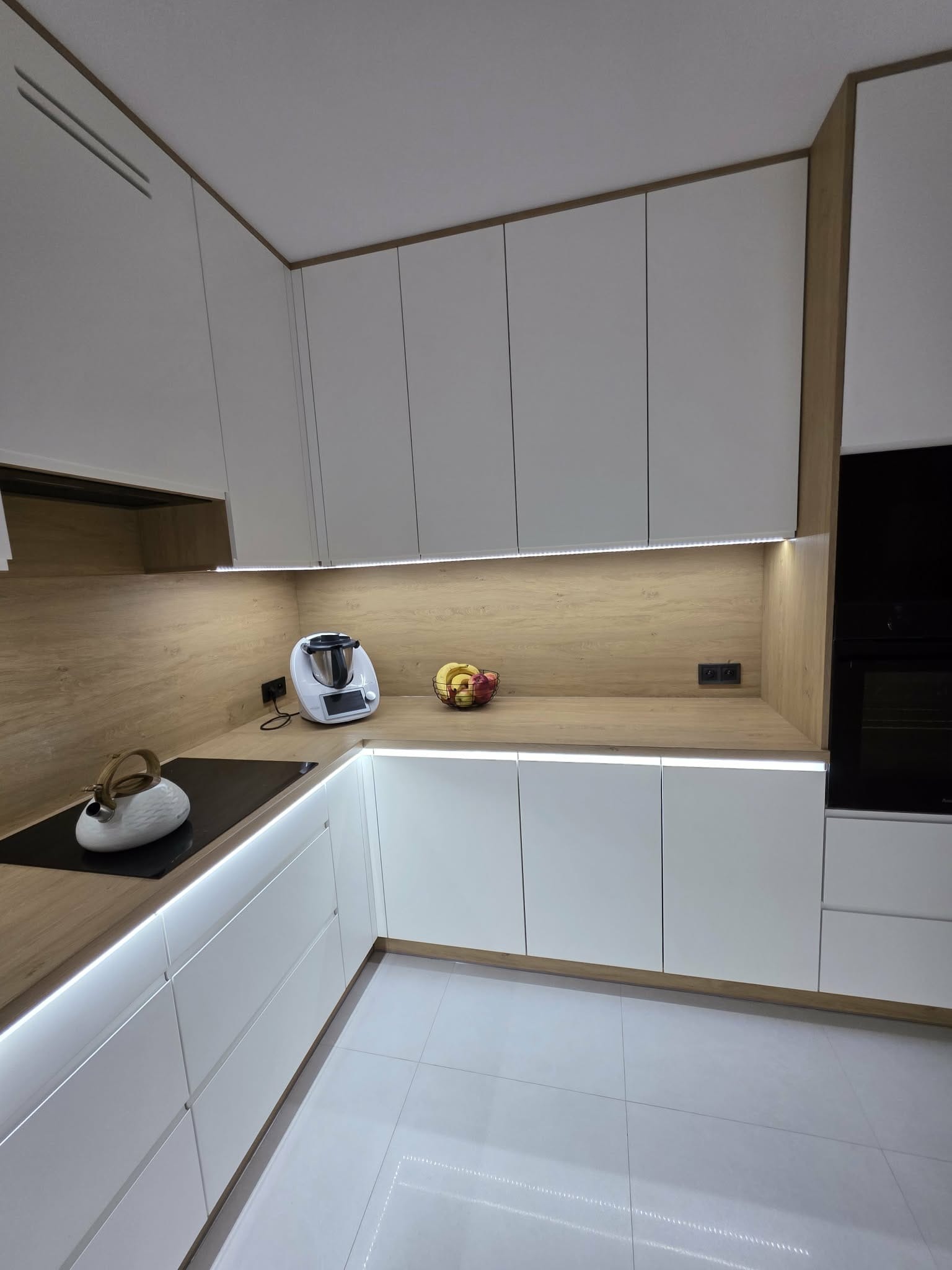 Nowoczesna kuchnia z białymi frontami i drewnianym blatem, oświetlona listwami LED. Na blacie Thermomix i misa z owocami. Minimalistyczny design, jasne wnętrze.
