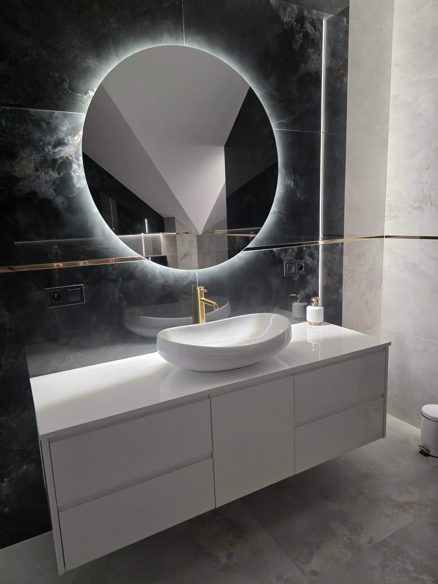 Nowoczesna łazienka z umywalką nablatową, szafką pod umywalkę w kolorze białym i okrągłym lustrem z podświetleniem LED. Marmurowe ściany i podłoga.