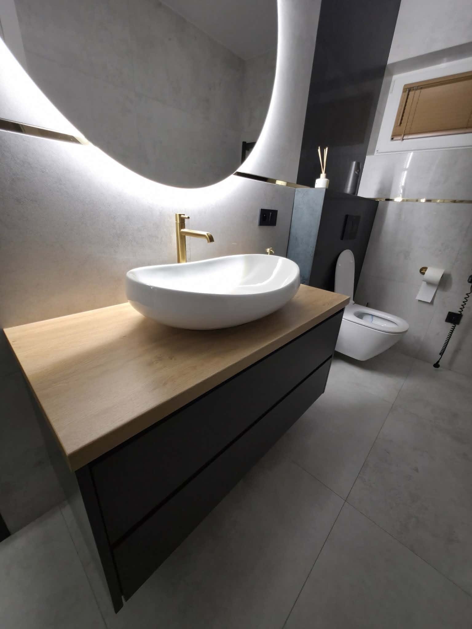 Nowoczesna łazienka z owalną umywalką nablatową, złotą armaturą i podświetlanym lustrem. Szafka pod umywalką w kolorze czarnym z drewnianym blatem.