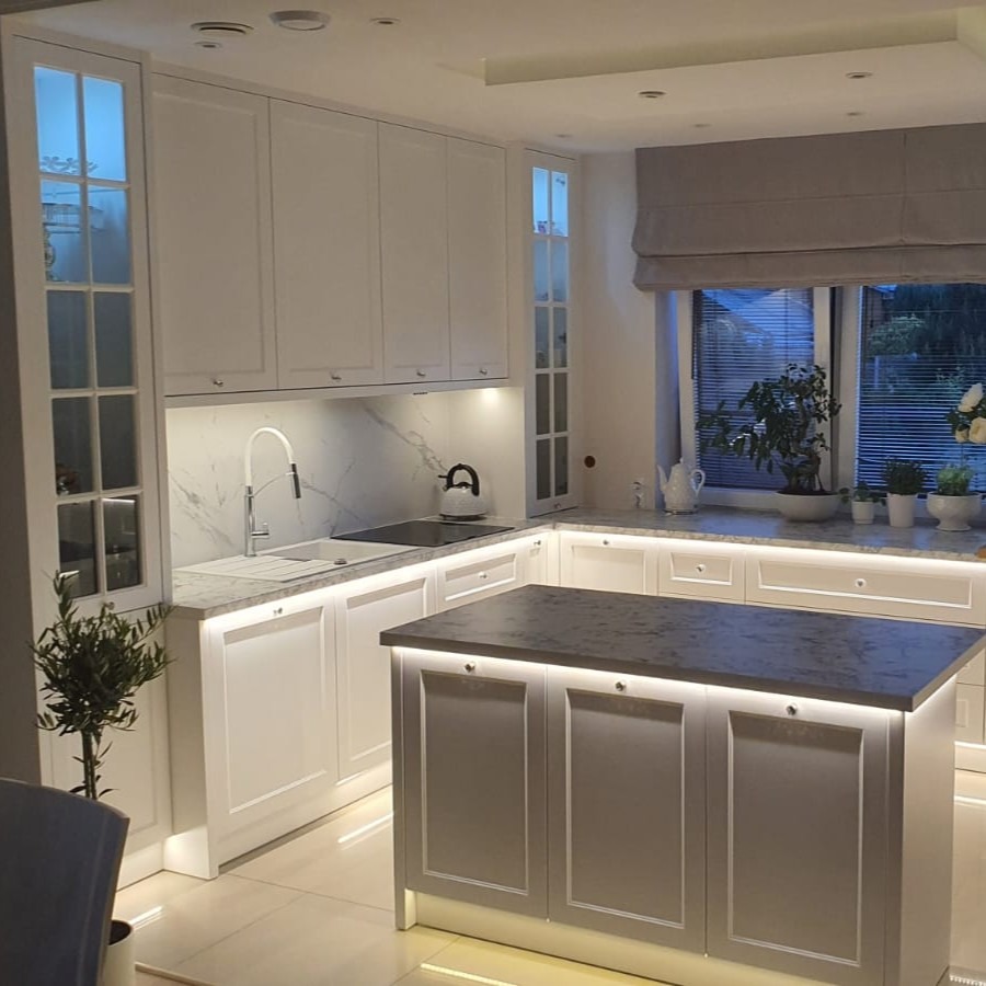 Jasna kuchnia z wyspą, oświetleniem LED pod szafkami i marmurowym blatem. Okno z roletą rzymską i roślinami. Nowoczesny design z elementami klasycznymi.