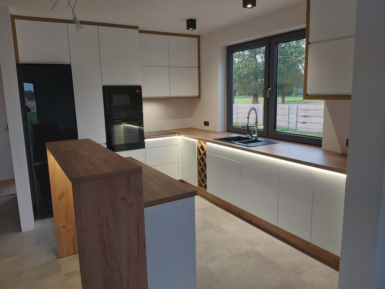 Nowoczesna kuchnia z białymi frontami, drewnianymi akcentami i oświetleniem LED pod szafkami. Widoczny zlew przy oknie i wyspa kuchenna z blatem w kolorze drewna.