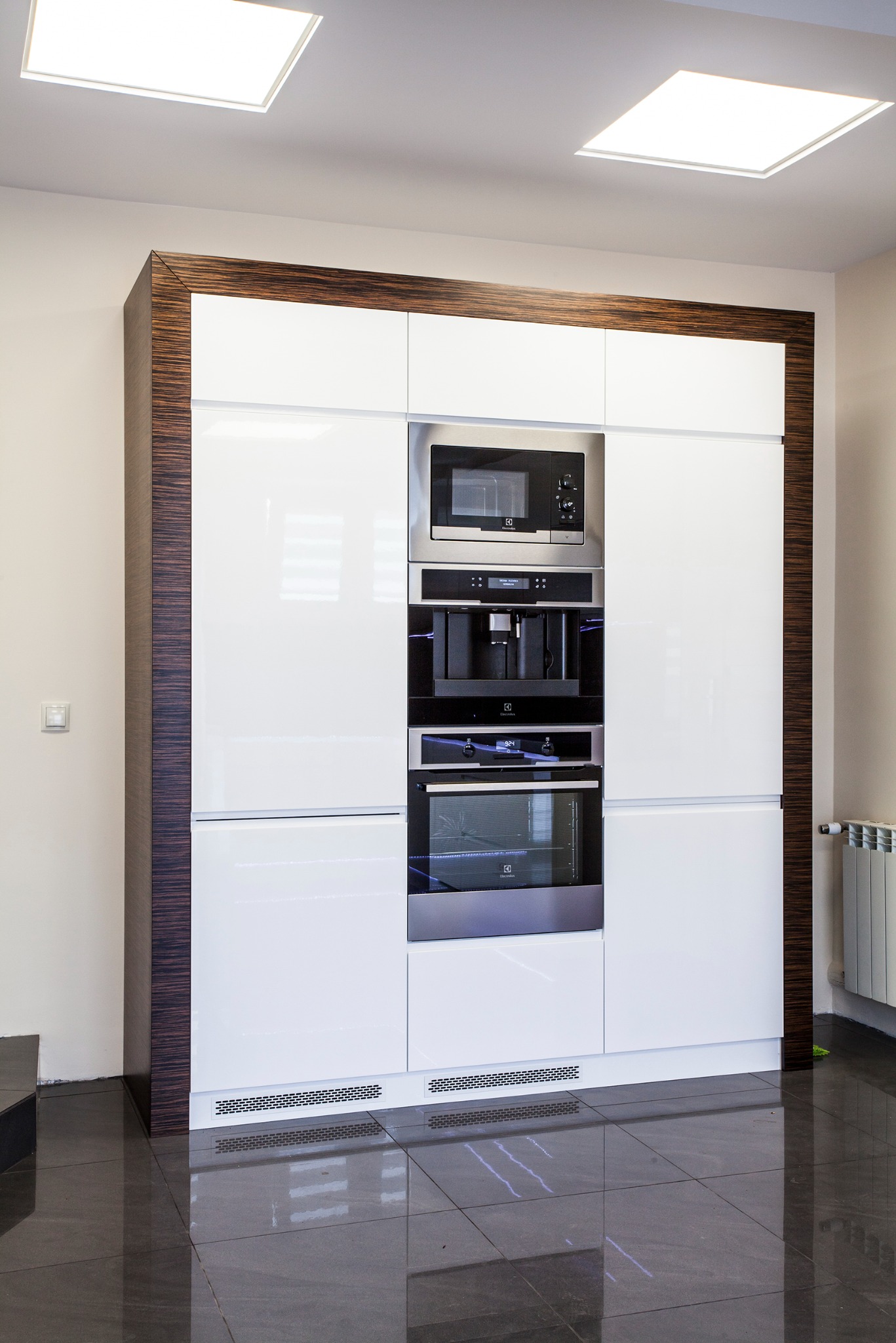Biała zabudowa kuchenna z wbudowanymi sprzętami Electrolux, obramowana ciemnym drewnem. Nowoczesny design, błyszczące fronty i ciemna podłoga.