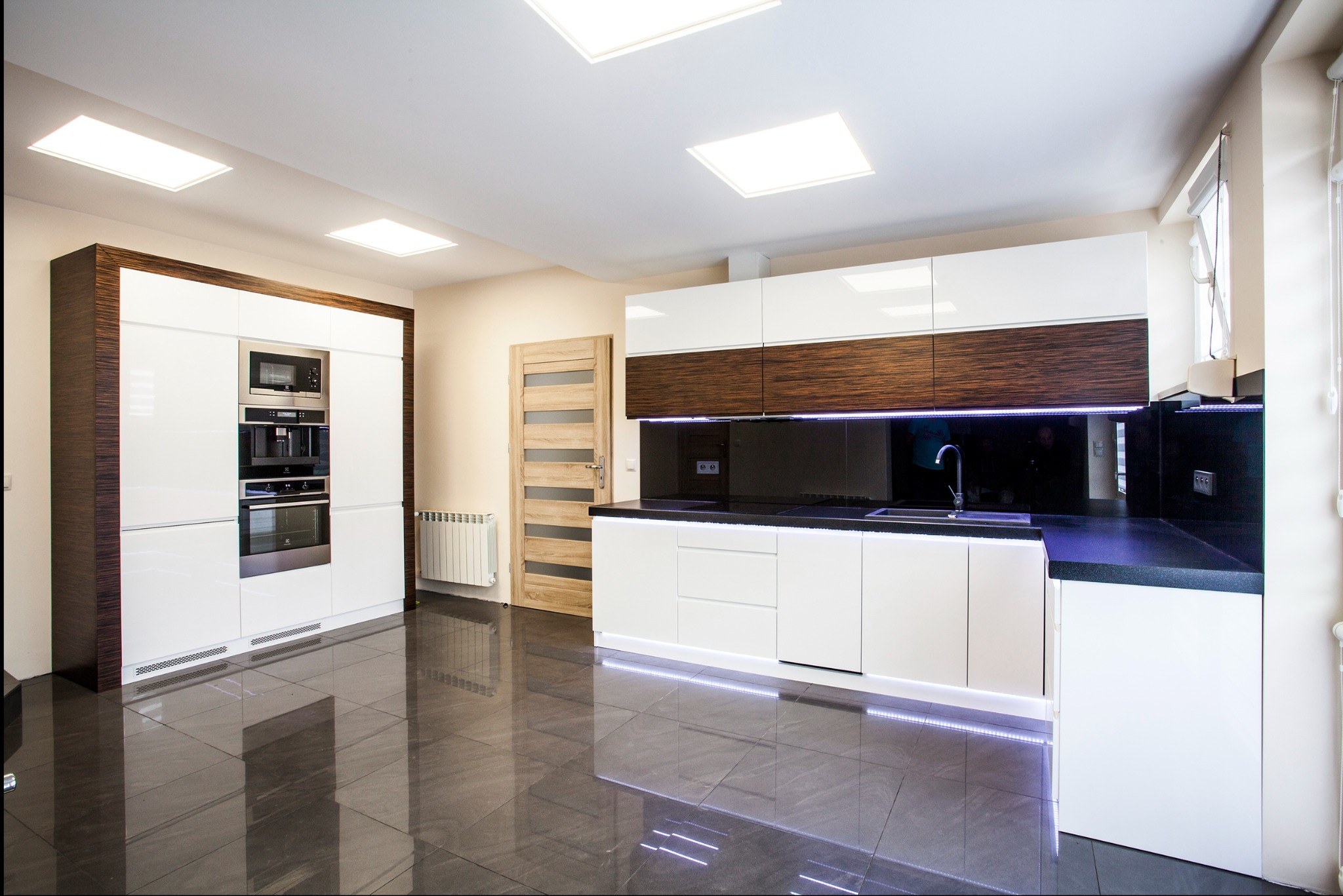 Nowoczesna kuchnia z białymi szafkami i drewnianymi akcentami, oświetleniem LED pod szafkami i na suficie, oraz błyszczącą, ciemną podłogą.