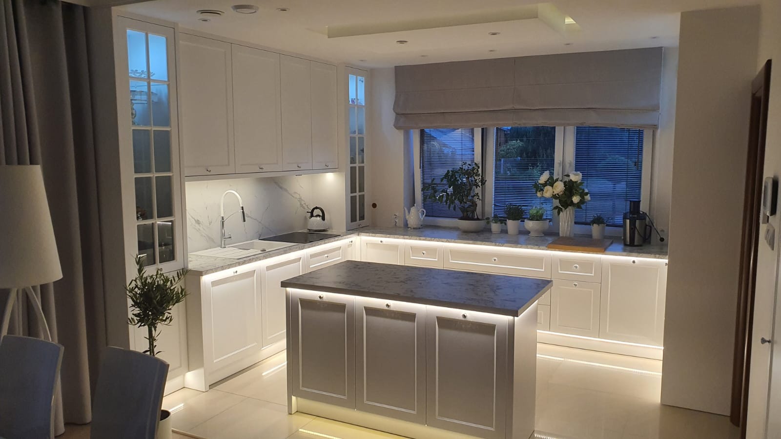 Jasna kuchnia z wyspą, oświetleniem LED i marmurowym blatem. Okno z roletą rzymską i roślinami. Nowoczesny design z elementami klasycznymi.