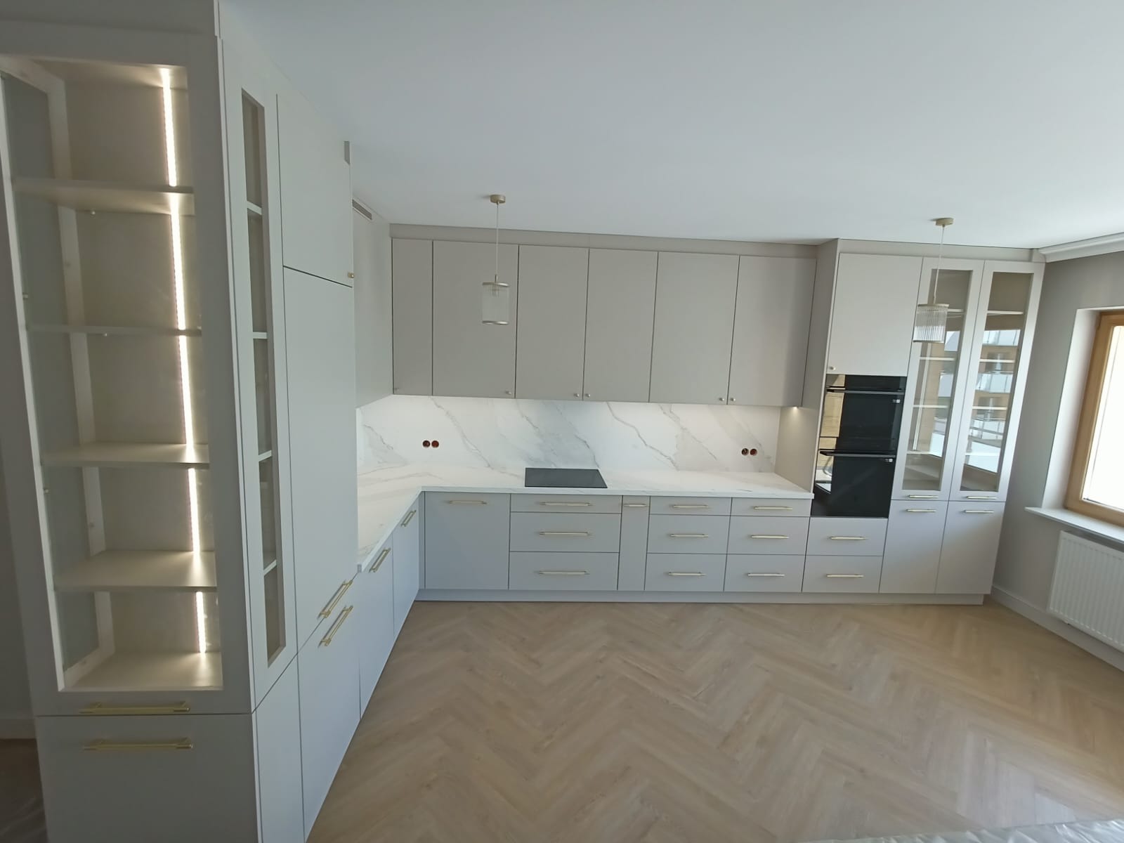 Jasna, nowoczesna kuchnia z marmurowym blatem i oświetleniem LED w witrynie. Ustawienie w kształcie litery L, parkiet w jodełkę. Minimalistyczne uchwyty.