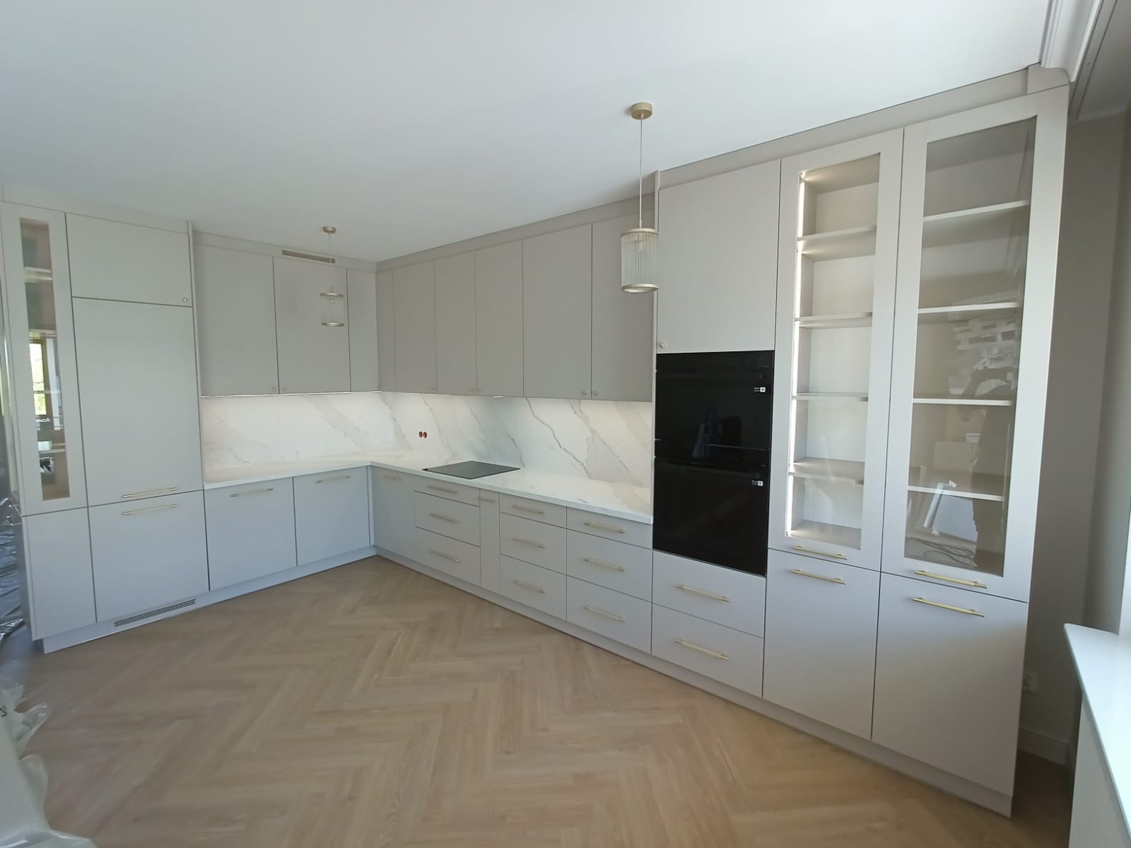 Jasna kuchnia z szarymi szafkami, marmurowym blatem i złotymi uchwytami. Na ścianie czarny piekarnik i szafki z przeszklonymi drzwiami. Parkiet w jodełkę na podłodze.