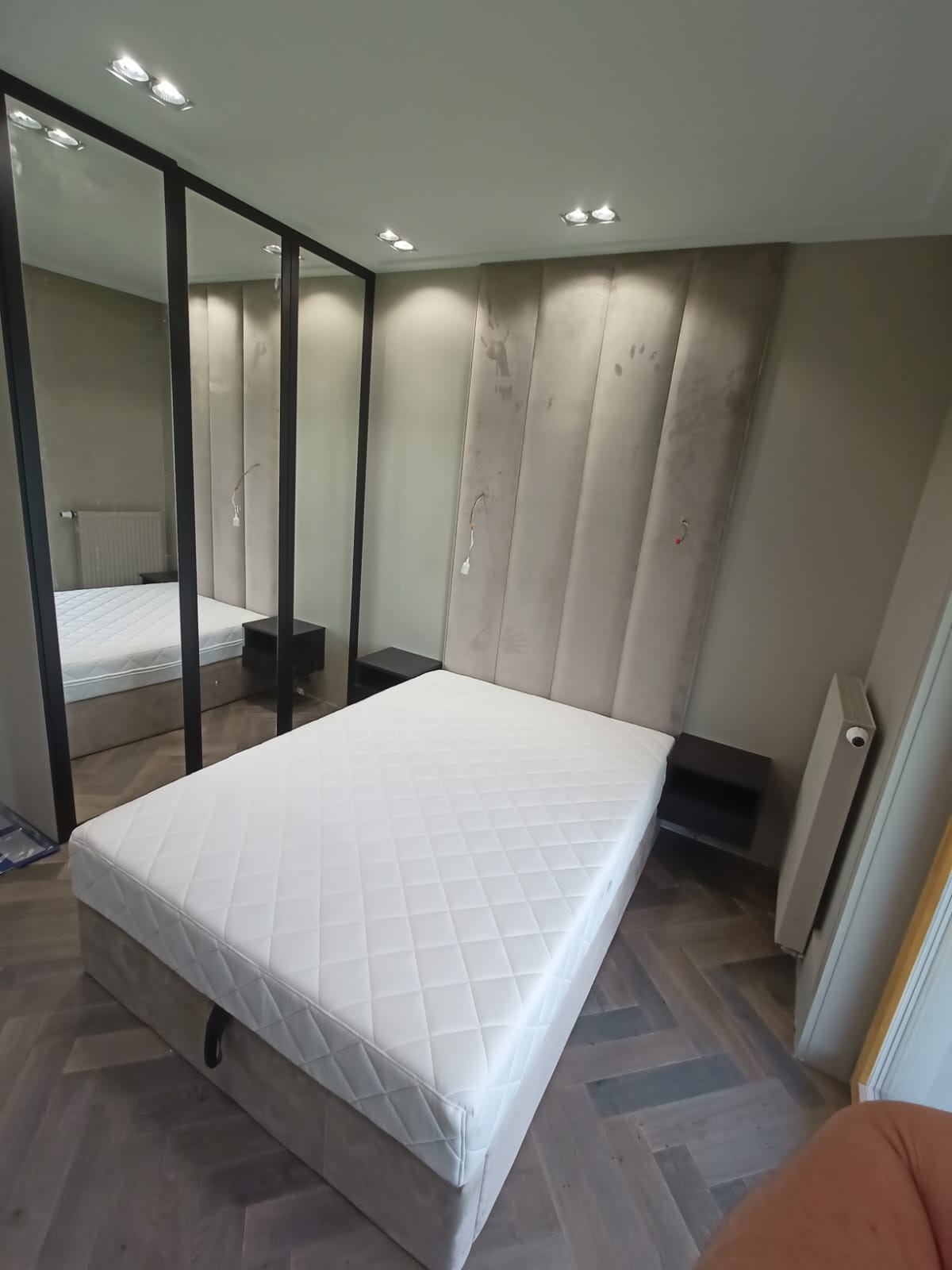 Nowoczesna sypialnia z łóżkiem tapicerowanym, panelem ściennym i szafą z lustrzanymi drzwiami w czarnych ramach. Parkiet ułożony w jodełkę. Minimalistyczny design.