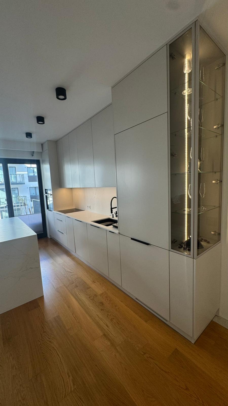 Nowoczesna kuchnia z białymi, matowymi frontami, czarną armaturą i przeszkloną witryną z oświetleniem LED. Parkiet na podłodze. Minimalistyczny design.