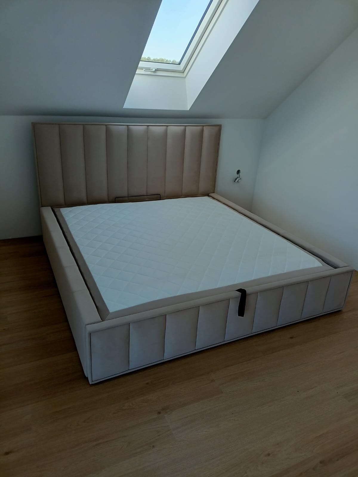 Beżowe, tapicerowane łóżko z pikowanym zagłówkiem w nowoczesnej sypialni z oknem dachowym. Widoczny fragment podłogi z paneli.