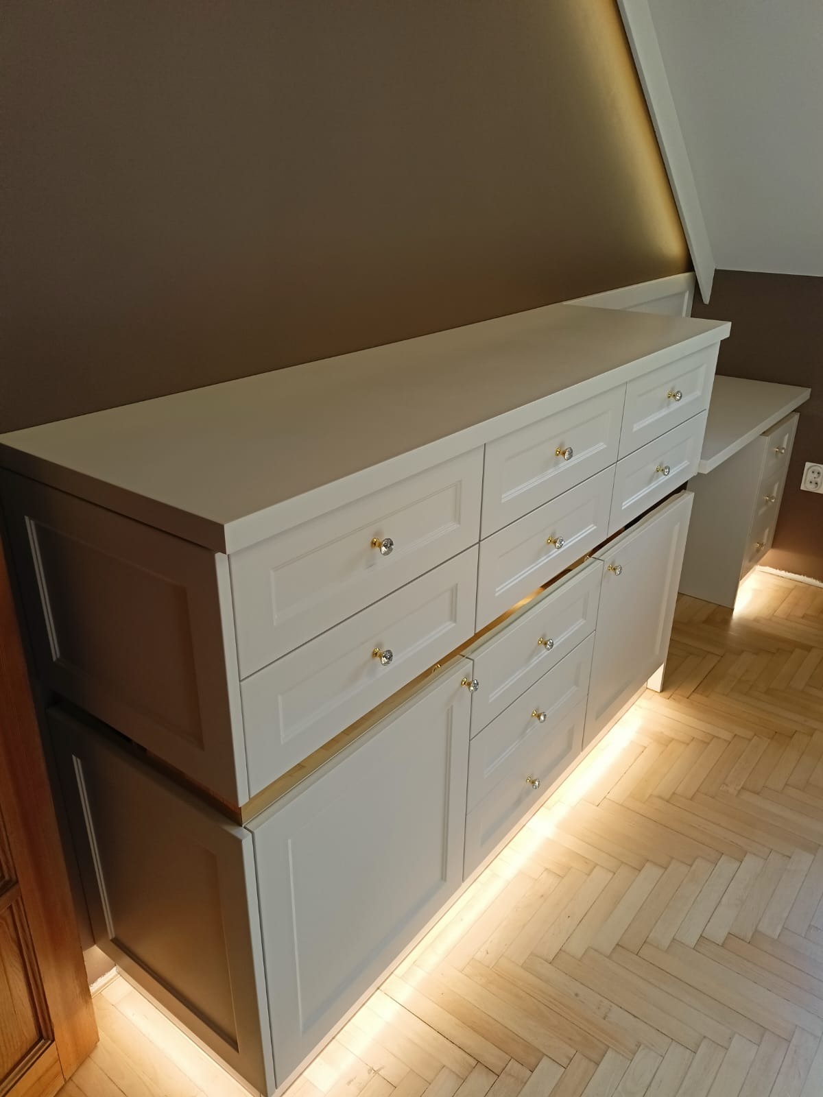 Biała szafa na wymiar z szufladami i oświetleniem LED, dopasowana do skosu poddasza. Widoczna drewniana podłoga i brązowa ściana.