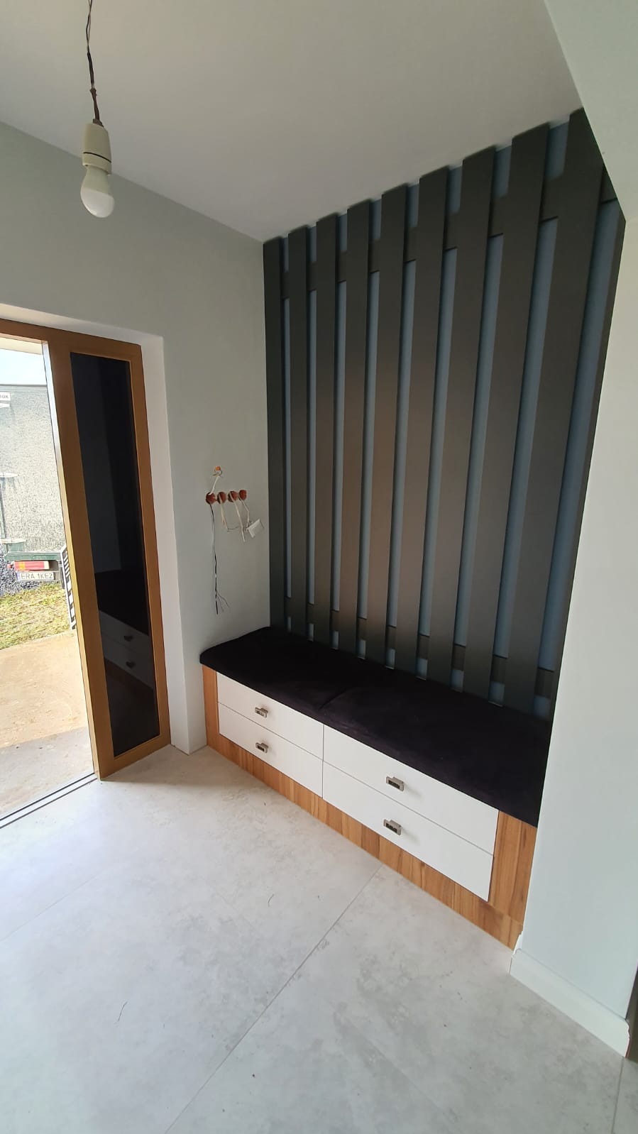 Nowoczesna zabudowa wnęki z siedziskiem i dekoracyjnymi panelami ściennymi w odcieniach szarości. Białe fronty szuflad z metalowymi uchwytami, drewniane wykończenie.