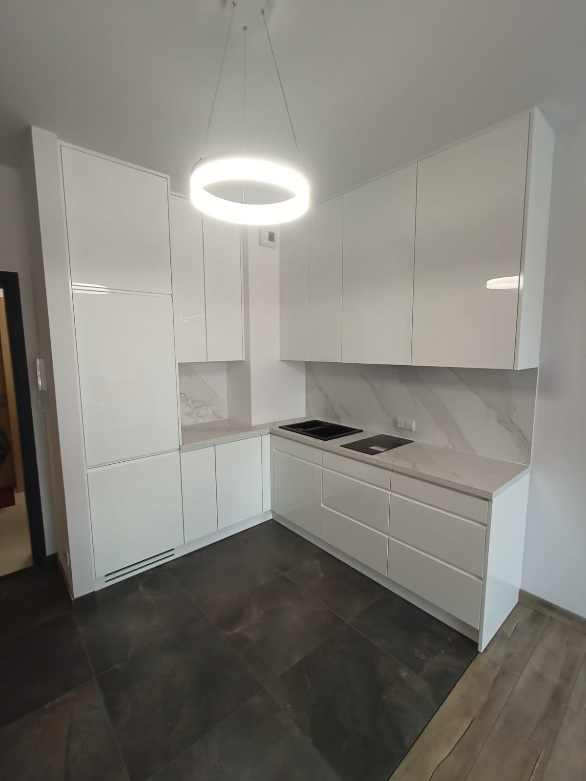 Biała, nowoczesna kuchnia z połyskiem, marmurowy backsplash, czarne płyty grzewcze i okrągła lampa sufitowa. Minimalistyczny design, ciemna podłoga.