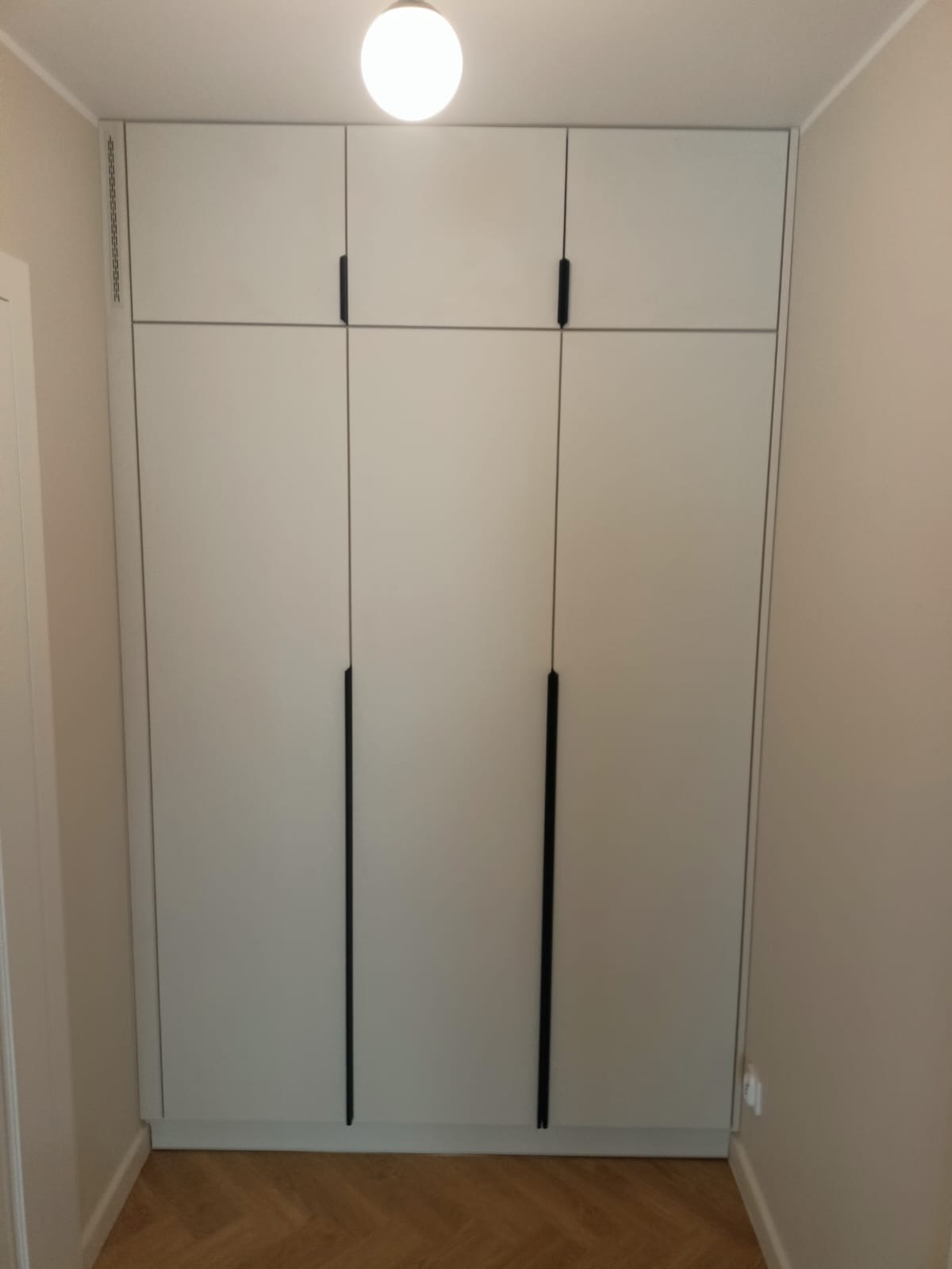 Biała szafa wnękowa z czarnymi, pionowymi uchwytami, dopasowana do wnęki w przedpokoju. Parkiet na podłodze, widoczny fragment ściany w neutralnym odcieniu.