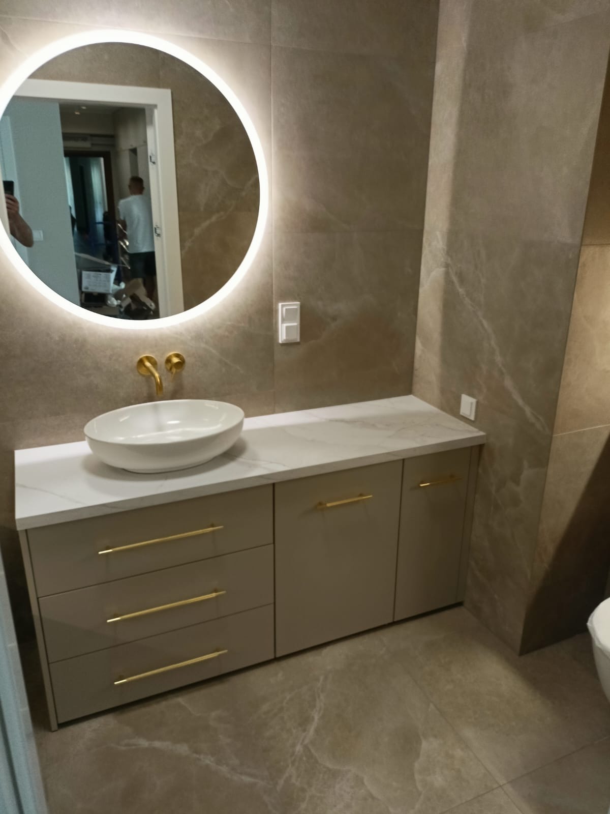 Nowoczesna łazienka z szafką na wymiar w odcieniach beżu, z białym blatem i złotymi uchwytami. Okrągłe lustro z podświetleniem LED. Płytki imitujące marmur.