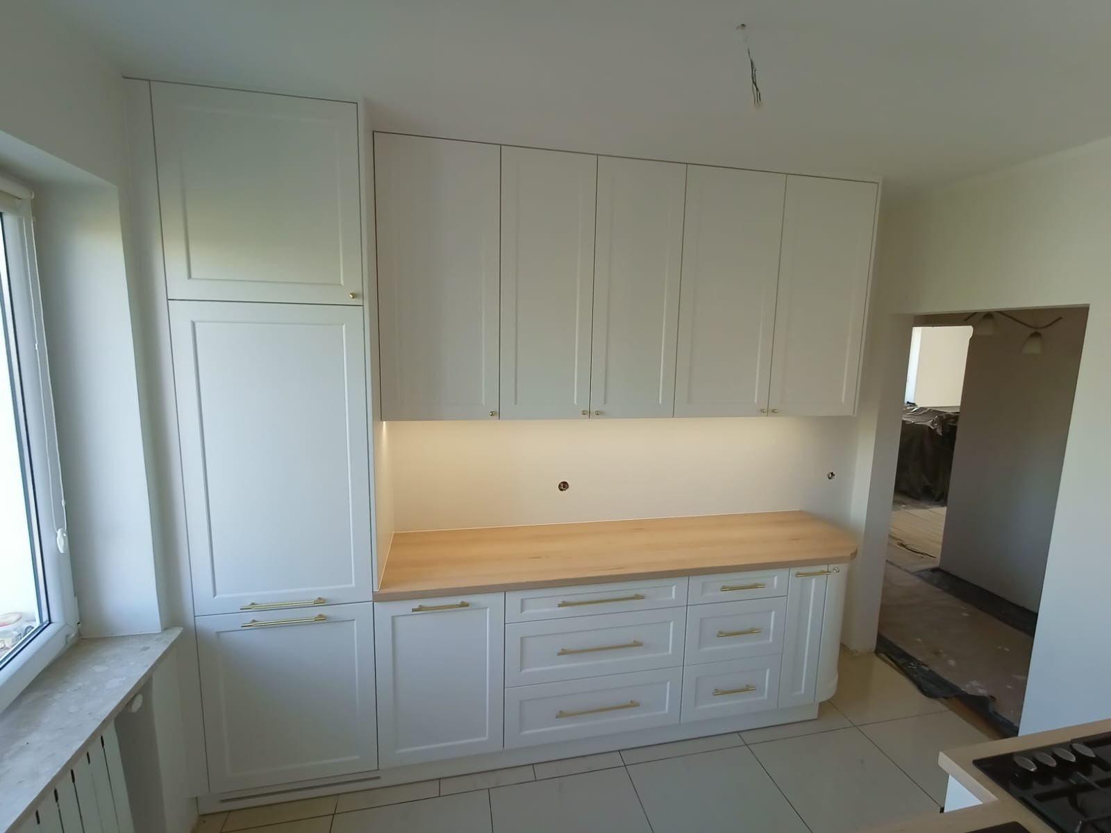Biała zabudowa kuchenna z drewnianym blatem i złotymi uchwytami, oświetlenie podszafkowe, widok na sąsiednie pomieszczenie w trakcie remontu.