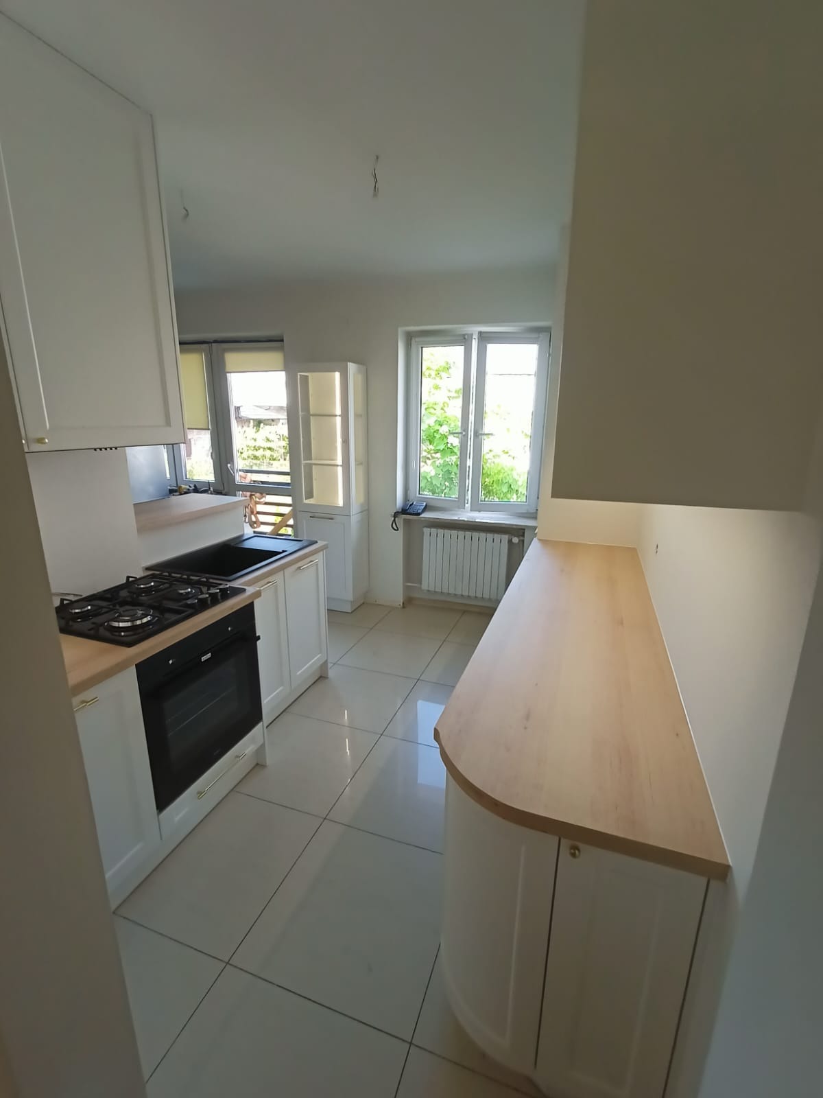 Jasna kuchnia z białymi szafkami i drewnianym blatem, widok z perspektywy wejścia, z oknem w tle. Minimalistyczny design, płytki podłogowe w odcieniach beżu.