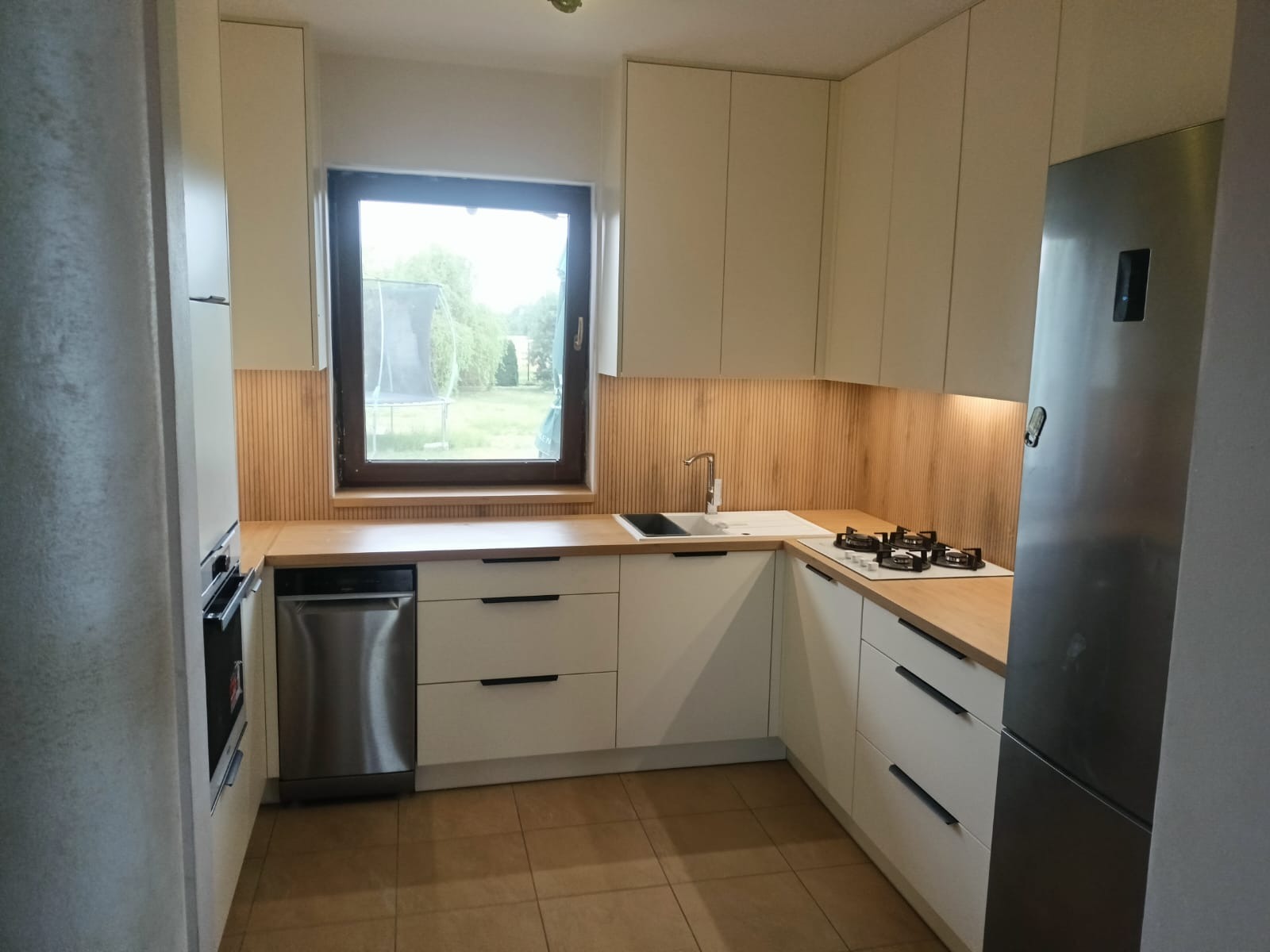 Jasna kuchnia z drewnianym akcentem na ścianie, widok na ogród przez okno. Minimalistyczne fronty szafek i blaty z naturalnego drewna tworzą przytulną atmosferę.