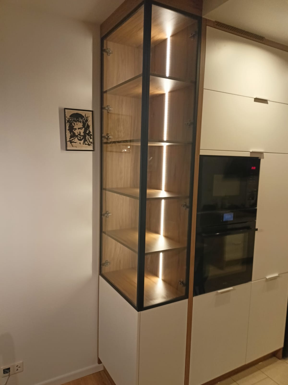 Nowoczesna, przeszklona witryna z oświetleniem LED, wkomponowana w zabudowę kuchenną z piekarnikiem. Na ścianie grafika w czarnej ramce.