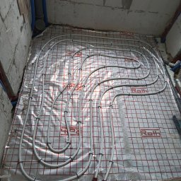 Jacek Godlewski - Ułożone rury ogrzewania podłogowego na folii izolacyjnej z siatką w surowym wnętrzu. Widoczne mocowania rur i fragmenty instalacji wodnej.