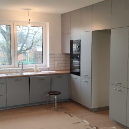 Jacek Godlewski - Nowoczesna kuchnia w szarościach z drewnianym blatem i białymi płytkami. Widoczny okap, okno z widokiem na drzewa i fragment zielonej ściany. Minimalistyczny design.