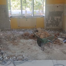 Jacek Godlewski - Demontaż płytek w kuchni: gruz na podłodze, taczka z resztkami, ściany częściowo skute, okno w tle. Etap przygotowawczy do remontu kuchni.
