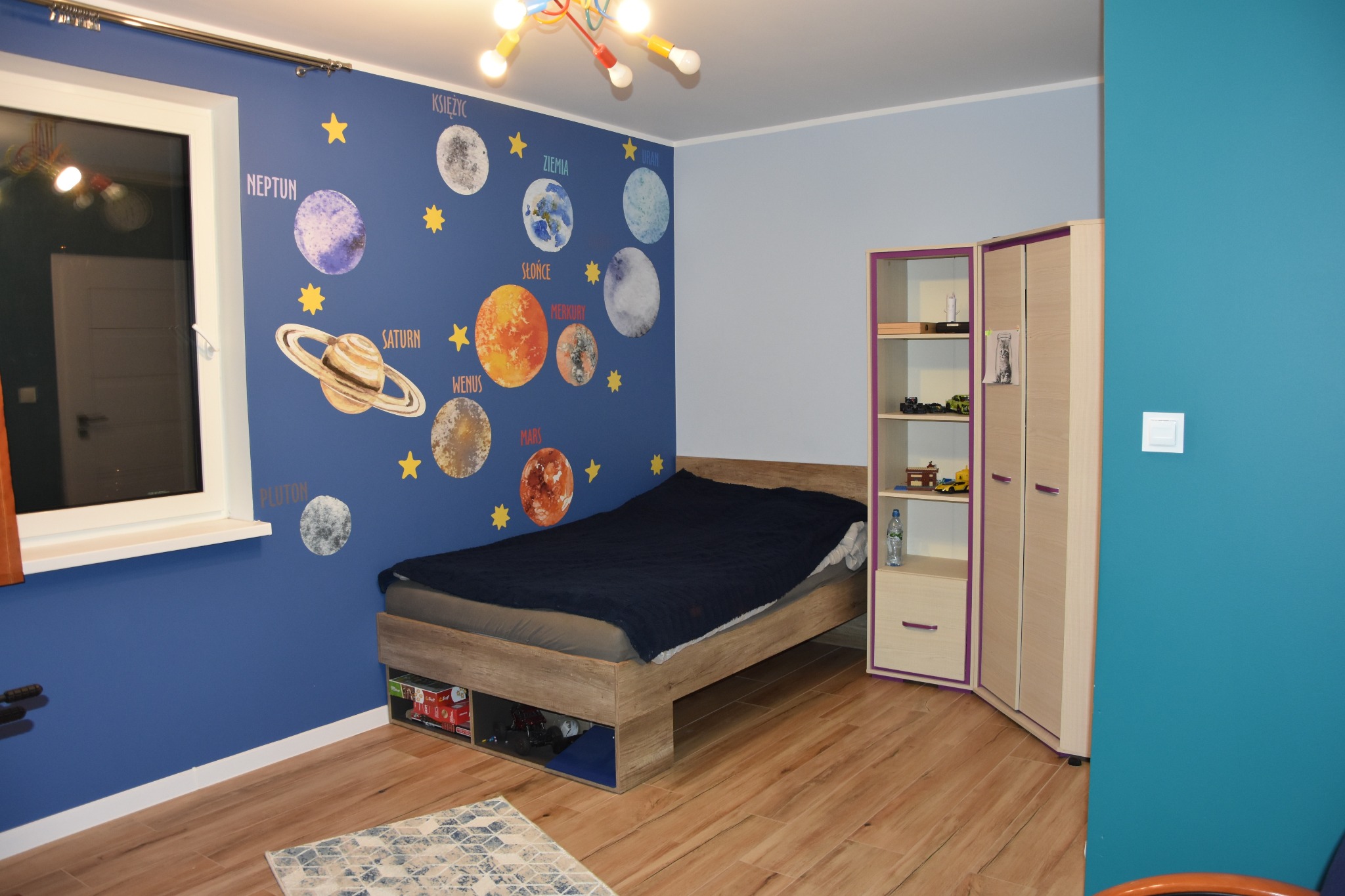 Pokój dziecięcy z motywem kosmosu: ściana w kolorze granatu z planetami, drewniane łóżko i szafa w odcieniach beżu i fioletu, drewniana podłoga i dywanik.