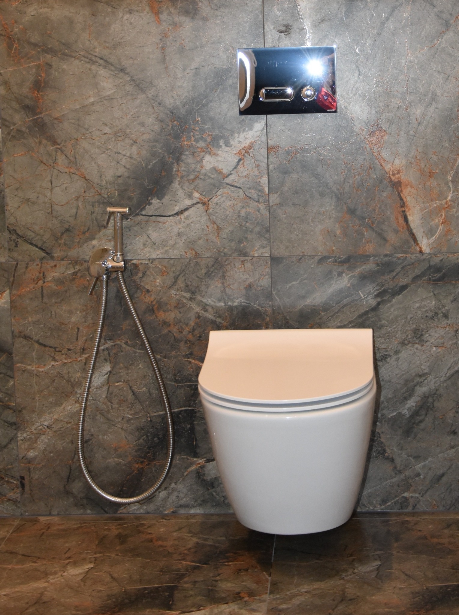 Nowoczesna toaleta podwieszana z bidetką i chromowanym przyciskiem spłukującym na tle płytek imitujących marmur w odcieniach szarości i brązu. Eleganckie wykończenie łazienki.